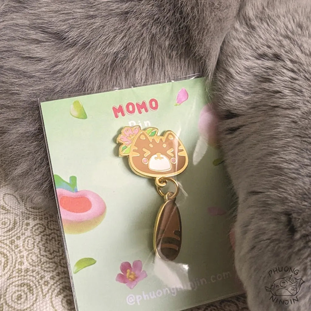 Momo cat enamel pin with golden edges. Packaged up next to the fluffy cat Daiki