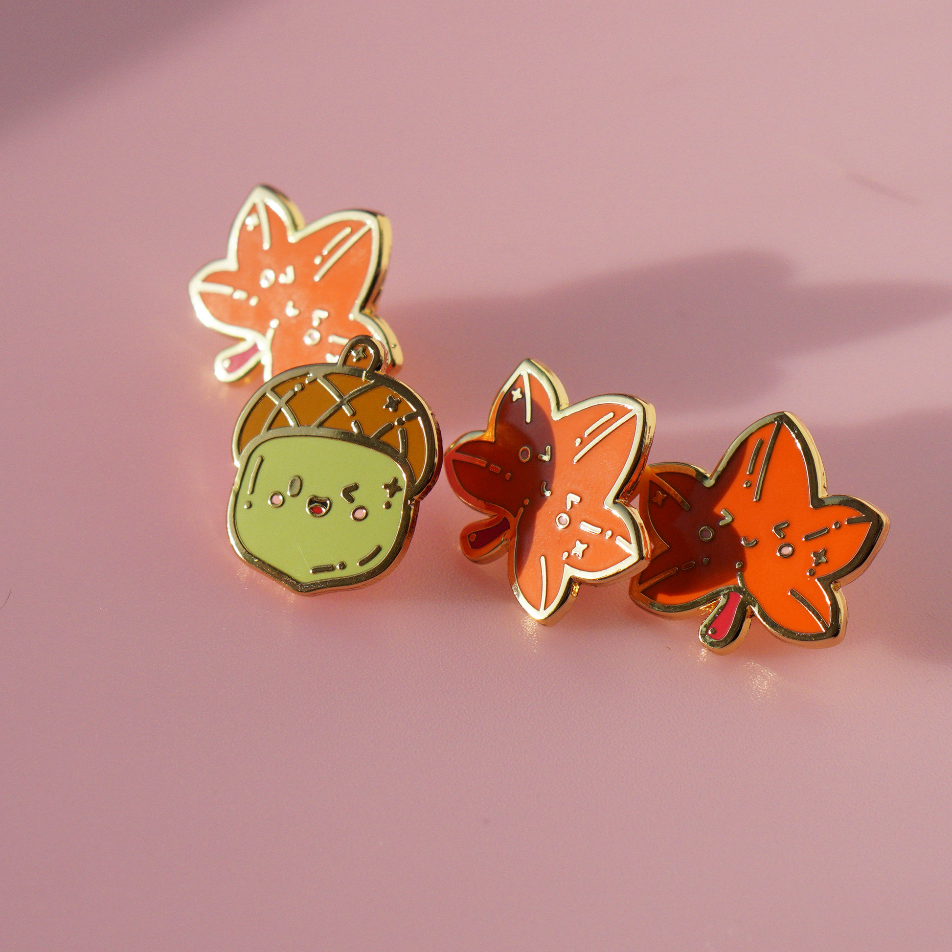 Green acorn enamel pin with golden outlines. Displayed here surrounded by 3 maple leaf enamel pins