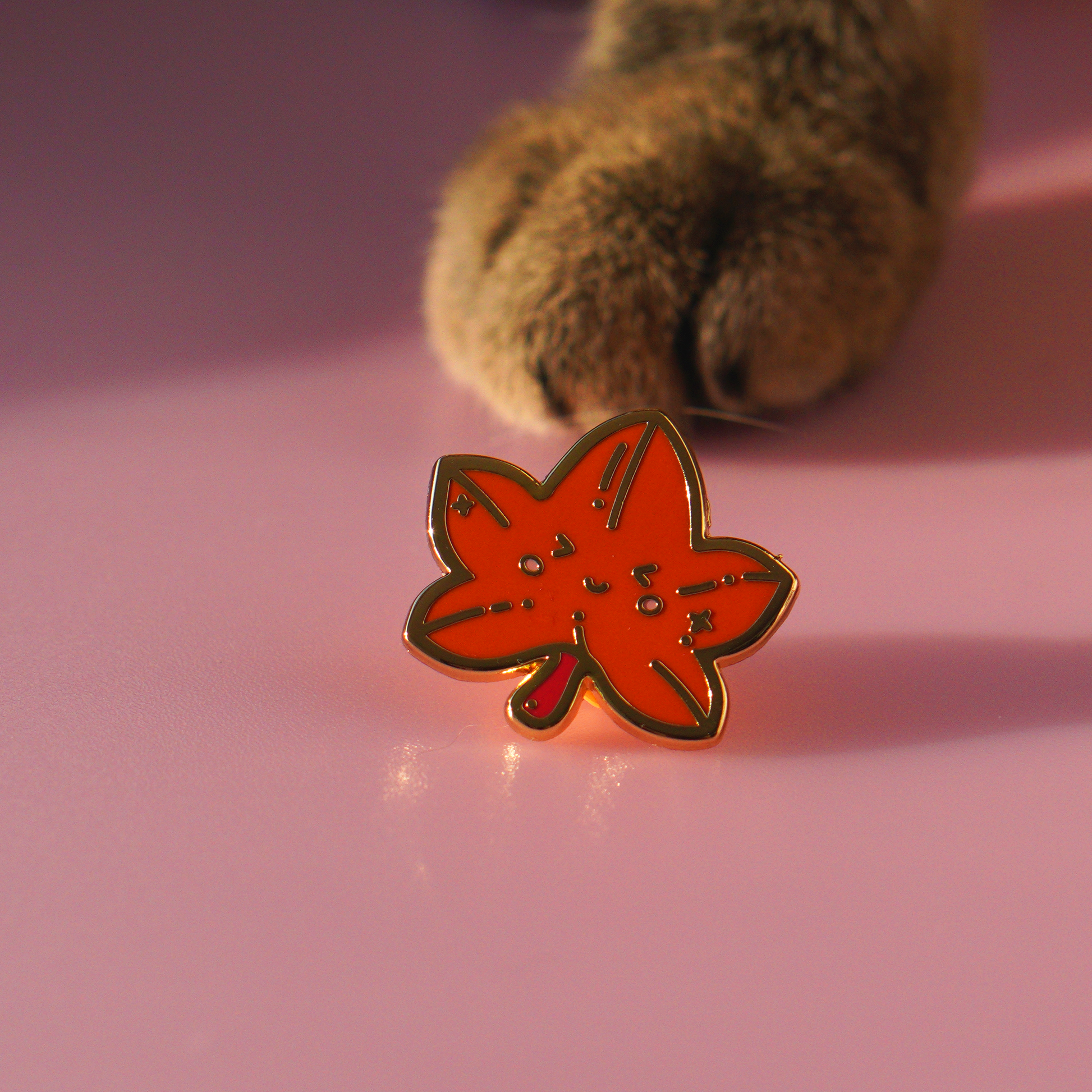 Maple leaf enamel pin with golden edges displayed next to momo her paw