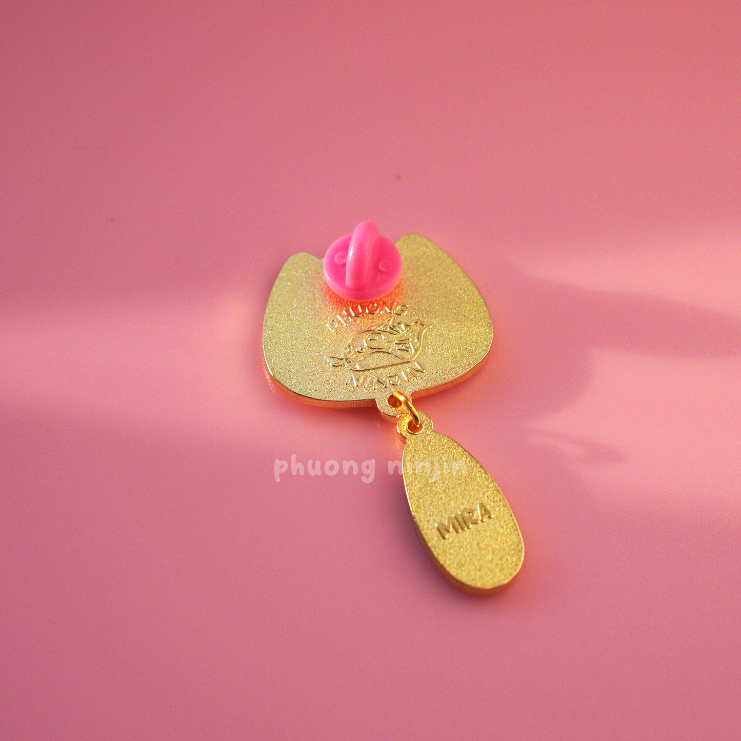 Mira cat enamel pin with golden edges. Displayed from the back, shown here the 'Phuong Ninjin' logo and the name 'Mira' embossed.