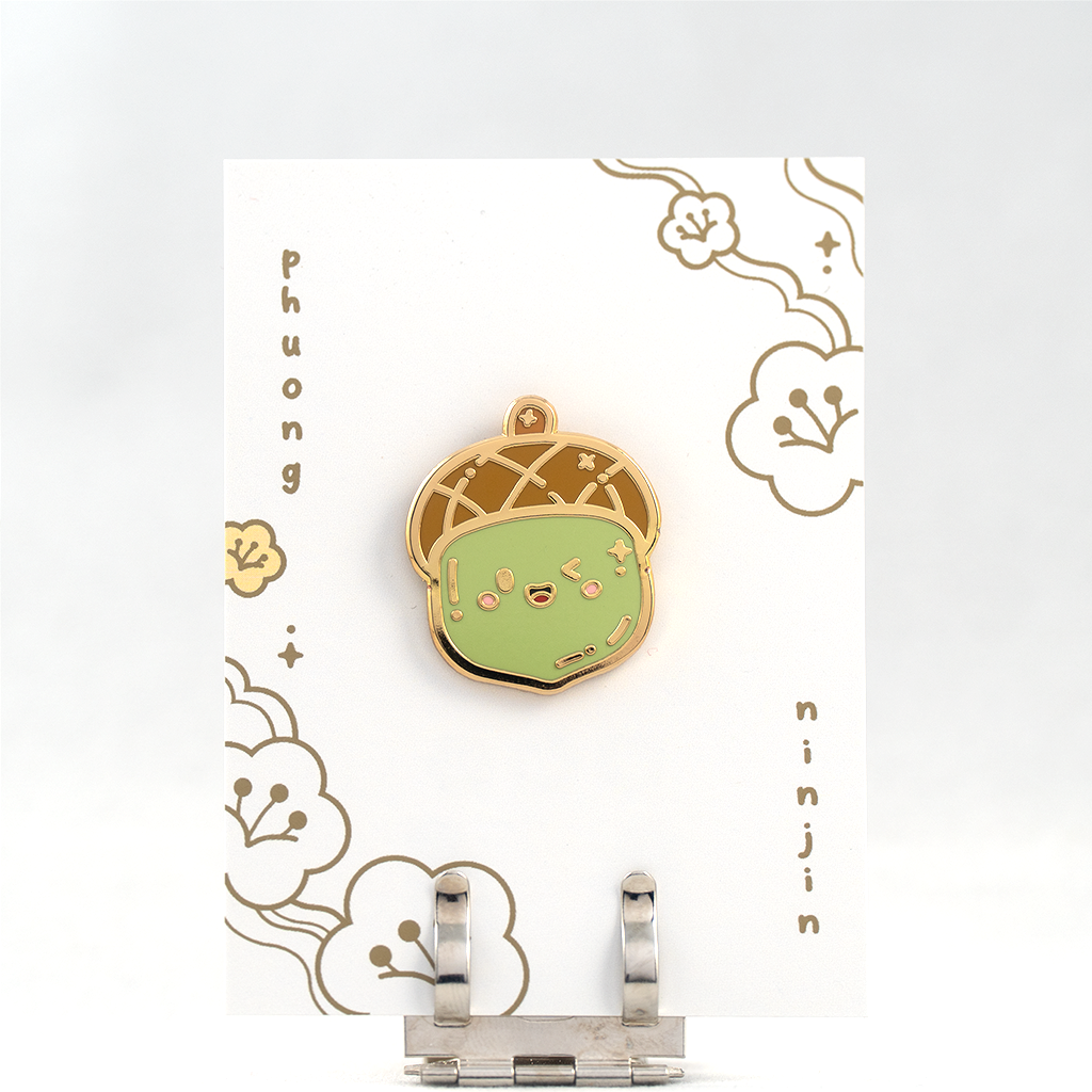 Green acorn enamel pin with golden outlines. Displayed here on the back card.