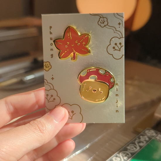 Autumn Mushroom enamel pin with golden edges displayed on our back card next to a maple leaf enamel pin video