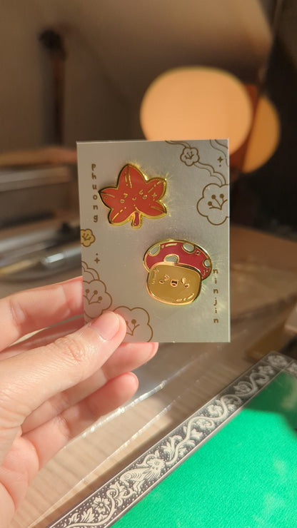 Autumn Mushroom enamel pin with golden edges displayed on our back card next to a maple leaf enamel pin video