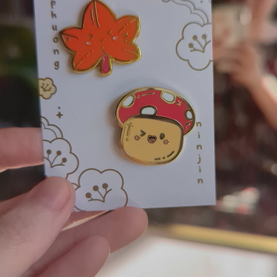 Maple leaf enamel pin with golden edges displayed along side our mushroom enamel pin on our back card, video.