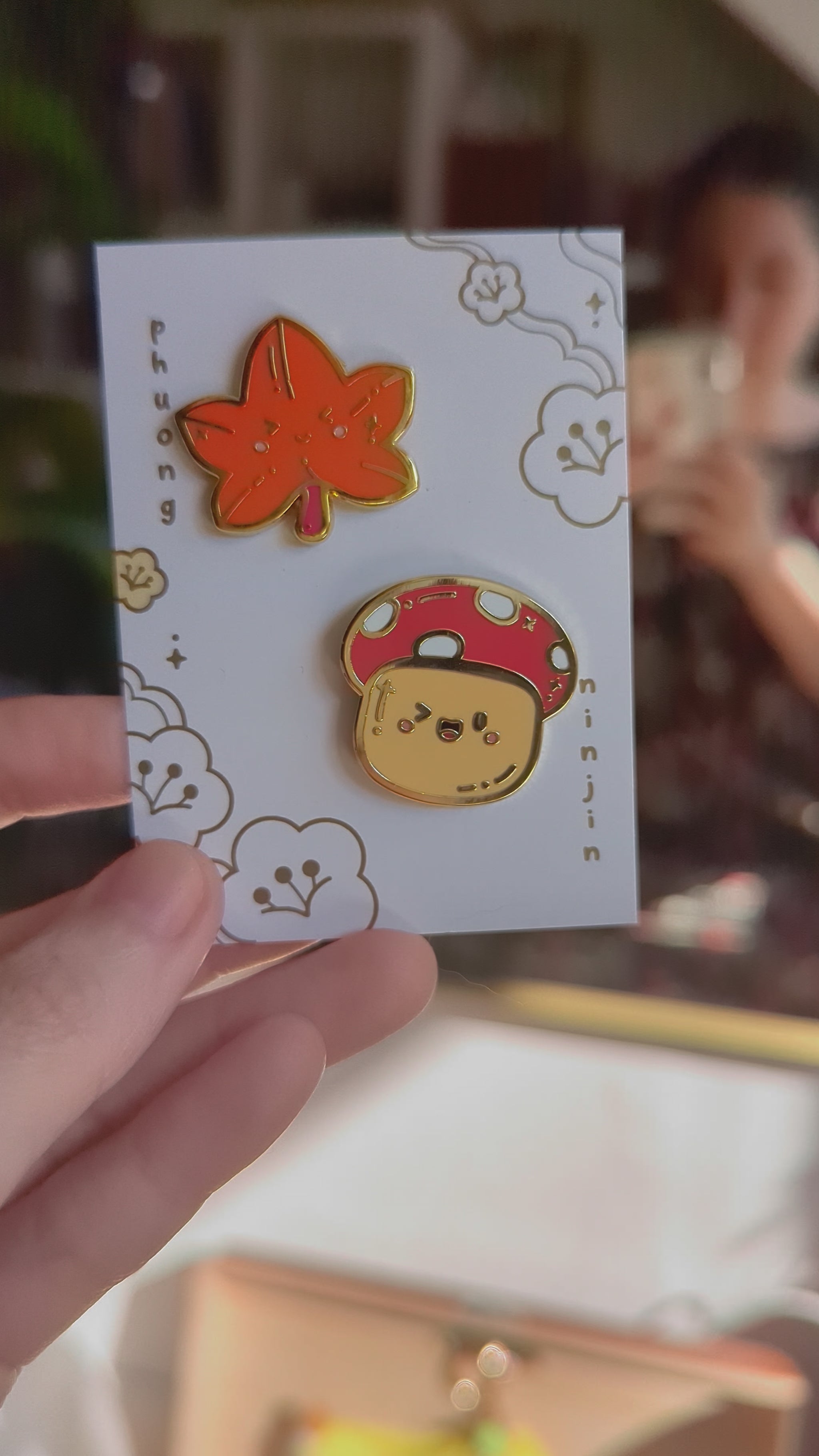 Maple leaf enamel pin with golden edges displayed along side our mushroom enamel pin on our back card, video.